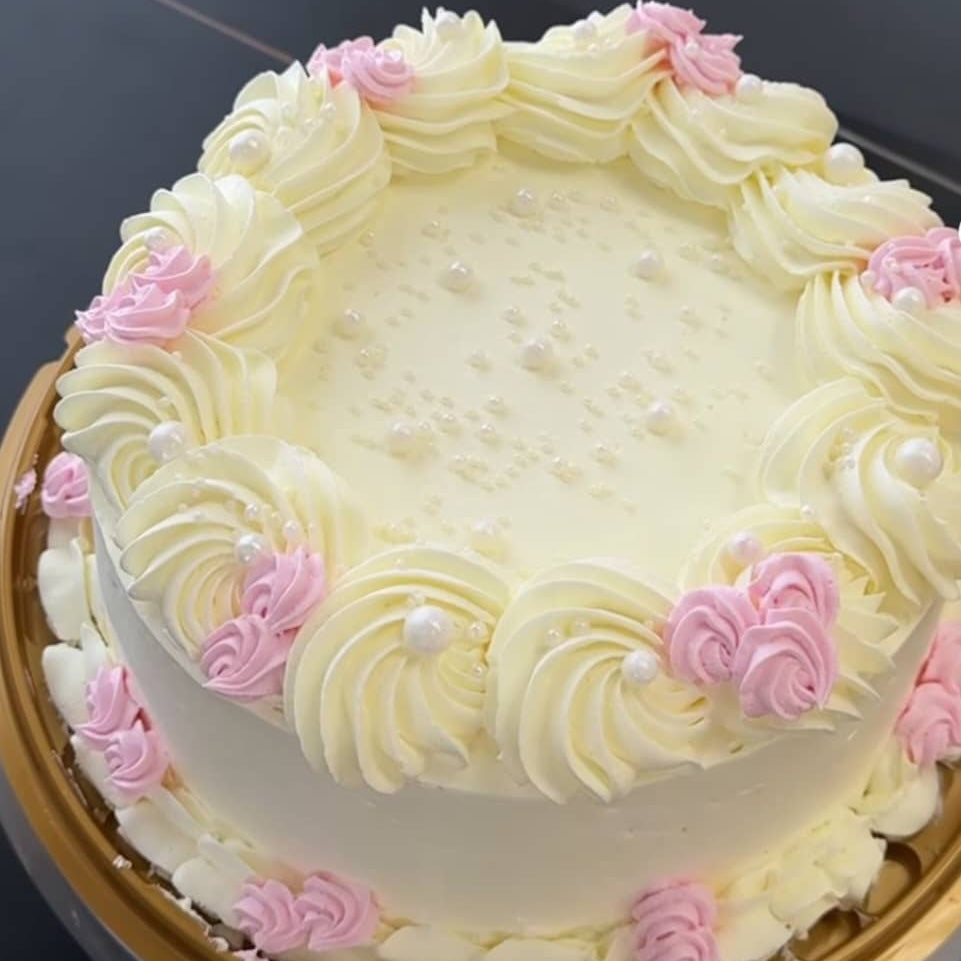 A round birthday cake decorated with white buttercream piped rosettes around the border, small pink rosettes, and white pearl-like sprinkles on top, sitting on a gold cake stand.