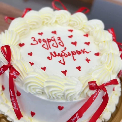 A round white birthday cake decorated with piped cream swirls, small red hearts, and red cursive writing on top, tied with red ribbon bows. The cake is photographed close up on a tray.